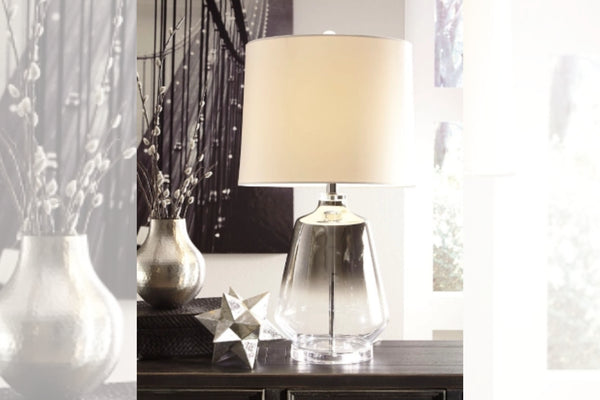 Lámpara decorativa con base metálica y pantalla blanca sobre mesa con elementos decorativos.