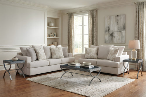 Juego de sala contemporáneo con sofá y loveseat tapizados en tela color marfil, cojines decorativos a juego, mesa de centro con estructura metálica y mesas auxiliares. Ambientado en una sala luminosa con alfombra clara y decoración en tonos neutros.