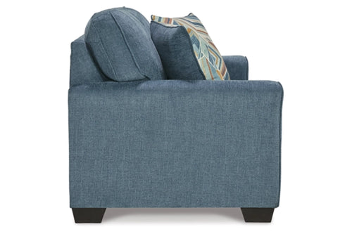 Vista lateral de loveseat de color azul con cojines sobre un fondo blanco. 