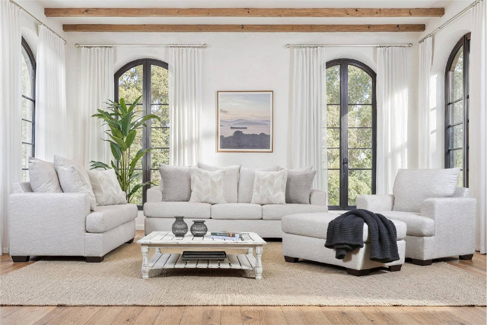 Sala luminosa con sofá, sillón y chaise longue tapizados en tela beige claro, decorados con cojines neutros. Incluye mesa de centro blanca, alfombra beige, grandes ventanas arqueadas y vigas de madera.
