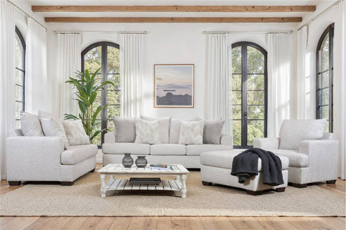 Sala luminosa con sofá, sillón y chaise longue tapizados en tela beige claro, decorados con cojines neutros. Incluye mesa de centro blanca, alfombra beige, grandes ventanas arqueadas y vigas de madera.