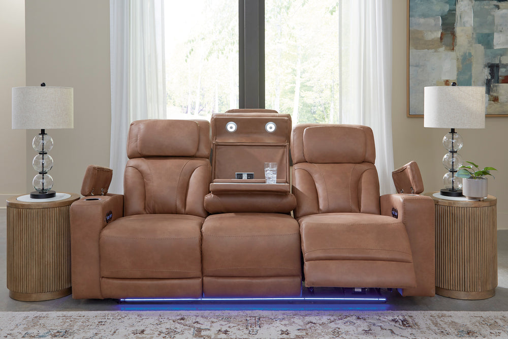 Loveseat reclinable en color café claro con consola central desplegable, puertos de carga integrados, iluminación LED azul en la base y reposapiés extendido en un asiento, ubicado entre mesas auxiliares con lámparas decorativas.
