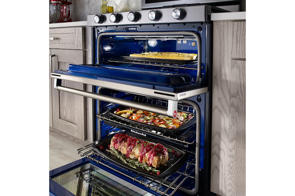 Horno doble empotrado con ambas puertas abiertas, que deja ver tres rejillas repletas de carnes asadas y productos horneados. El horno tiene un interior azul oscuro, exterior de acero inoxidable y mandos de control en el panel superior, integrado en un mueble de cocina.