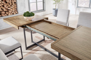 Mesa de comedor extensible con acabado en madera clara y estructura metálica negra. La imagen muestra el mecanismo de expansión con panel central desplegable, rodeada de sillas tapizadas en gris claro en un ambiente moderno y luminoso.
