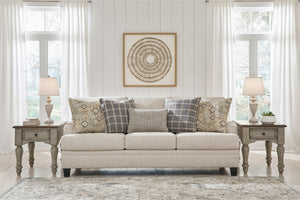 Sofá beige con cojines estampados en una habitación con paredes blancas, grandes ventanales y lámparas decorativas.