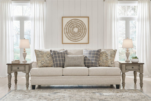 Sofá beige con cojines estampados en una habitación con paredes blancas, grandes ventanales y lámparas decorativas.