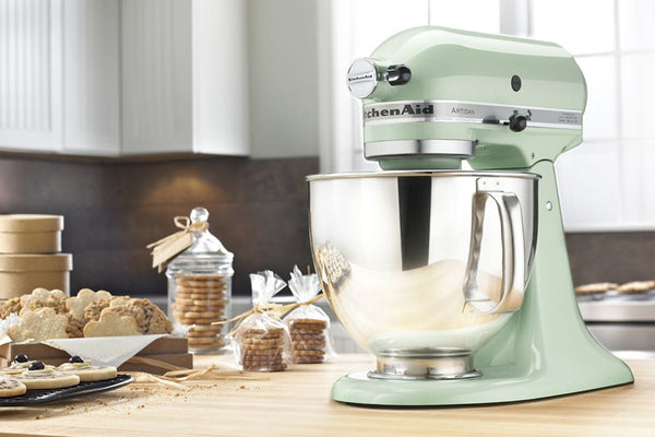 Una batidora KitchenAid de color verde salvia pálido se encuentra sobre una encimera de madera en una cocina, con galletas, tarros e ingredientes para hornear dispuestos al fondo.