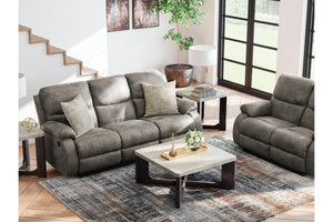 Un sofá reclinable tapizado en gris y un loveseat a juego se disponen en una luminosa sala de estar moderna con una alfombra estampada, una mesa de centro, mesas auxiliares y grandes ventanales.