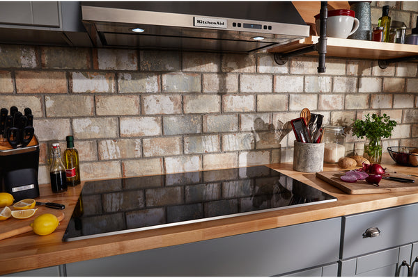 Un protector contra salpicaduras de cocina hecho de azulejos rectangulares de imitación de ladrillo de color beige claro y gris, montado detrás de una placa de cocción de vidrio negro, con encimeras de madera y artículos de cocina que incluyen limones, botellas, utensilios y una tabla de cortar.