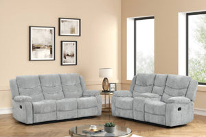 Juego de sala reclinable en tela gris claro compuesto por sofá y loveseat. Presentado en una sala luminosa con ventanas amplias, mesa de centro redonda y decoración moderna en tonos neutros para un ambiente acogedor.