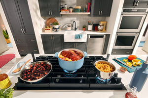Una escena de cocina moderna que muestra una placa de gas negra con una olla holandesa azul claro, una olla de acero inoxidable con arroz y dos sartenes con frijoles sobre la estufa. Al fondo se aprecian gabinetes gris oscuro, un fregadero blanco estilo granja y electrodomésticos empotrados de acero inoxidable.