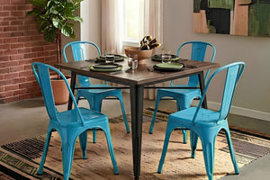 Mesa de comedor cuadrada con cubierta de madera oscura y estructura metálica negra, acompañada por cuatro sillas metálicas color azul turquesa de estilo industrial. El conjunto está sobre una alfombra decorativa en un comedor moderno con detalles naturales y luz ambiental.