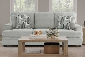 Sofá tapizado en tela gris claro, con cojines decorativos de patrón geométrico en tonos neutros. Presentado en una sala luminosa con mesa de centro de madera, arreglos florales y ventanas amplias al fondo.