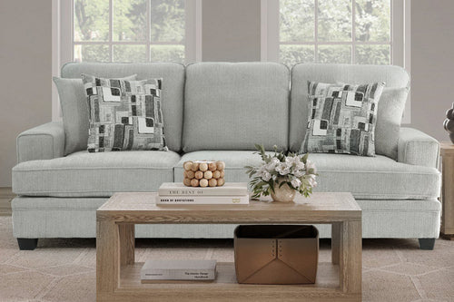Sofá tapizado en tela gris claro, con cojines decorativos de patrón geométrico en tonos neutros. Presentado en una sala luminosa con mesa de centro de madera, arreglos florales y ventanas amplias al fondo.