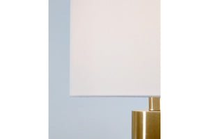 Lámpara de mesa con pantalla blanca y base dorada sobre una pared azul claro.