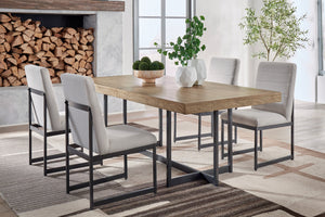 Comedor moderno con mesa rectangular de madera clara y estructura metálica negra, acompañado de cuatro sillas tapizadas en gris claro. Ambientado en un espacio luminoso con alfombra de diseño geométrico, plantas decorativas y chimenea con leña apilada.