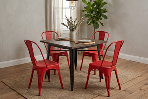 Mesa de comedor cuadrada con cubierta de madera oscura y estructura metálica negra, acompañada por cuatro sillas rojas de estilo industrial. El conjunto está sobre una alfombra tejida en un comedor moderno con planta decorativa y luz natural.