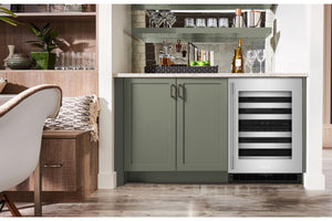 Una nevera para vinos empotrado bajo la encimera, con frontal de acero inoxidable y puerta de cristal, integrado en un moderno mueble de cocina con puertas de color verde salvia y una encimera clara.