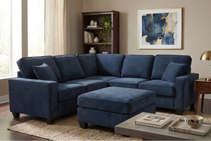 Sala de estar con sofá seccional en forma de L tapizado en tela azul y otomán a juego. El conjunto incluye cojines de respaldo y decorativos, y está ambientado en un espacio moderno con mesa de centro, librero, lámpara y arte de pared.