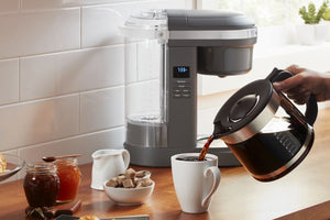 Una cafetera de sobremesa en una cocina luminosa, con una mano vertiendo agua caliente de una tetera negra de acero inoxidable en una taza blanca. La cafetera tiene un depósito de agua transparente y un cuerpo gris oscuro y plateado con una pequeña pantalla digital.