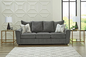 Sofá gris en sala con diseño geométrico de pared y grandes ventanales.