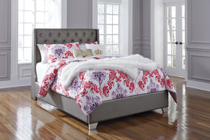 Cama con edredón estampado de flores y cabecero gris en una habitación neutral con ventanas. 