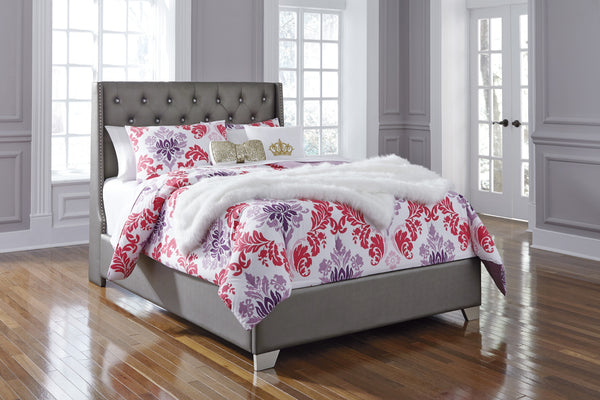 Cama con edredón estampado de flores y cabecero gris en una habitación neutral con ventanas. 