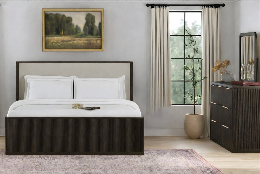 Dormitorio contemporáneo con cama de madera oscura y cabecera tapizada en tono claro, ropa de cama blanca, cómoda con espejo a juego, ventana con cortinas beige, planta decorativa y cuadro de paisaje sobre la cabecera.