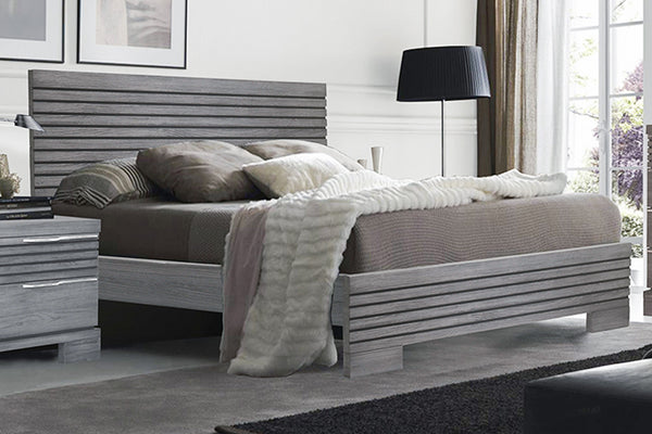 Grey, wooden bed with beige bedding in a room setting with a window to the right and decorate items. 