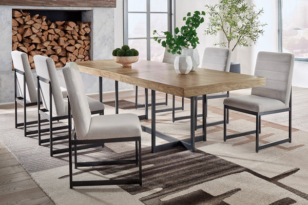 Juego de comedor contemporáneo con mesa rectangular de acabado madera clara y seis sillas tapizadas en gris claro con estructura metálica negra. Ambientado en un espacio luminoso con alfombra geométrica, plantas decorativas y chimenea con leña apilada.