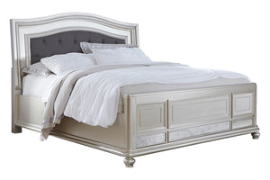 Cama tapizada y plateada con acolchado blanco y con cabecera gris oscura sobre un fondo blanco. 