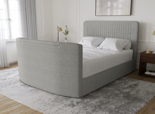 Una cama moderna tapizada con cabecero curvo y piecero a juego en tela gris claro, vestida con ropa de cama blanca y situada en un dormitorio luminoso con alfombra gris, suelo de madera y mesita de noche.
