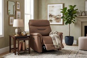 Sillón reclinable tapizado en color marrón con respaldo alto acolchado, reposapiés extendido y apoyabrazos amplios. Presentado en una sala acogedora junto a una mesa auxiliar, lámpara decorativa, planta en maceta y arte de pared en tonos neutros.