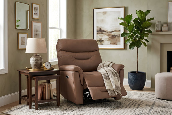 Sillón reclinable tapizado en color marrón con respaldo alto acolchado, reposapiés extendido y apoyabrazos amplios. Presentado en una sala acogedora junto a una mesa auxiliar, lámpara decorativa, planta en maceta y arte de pared en tonos neutros.