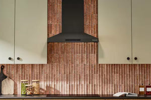 Una escena de cocina moderna que presenta una campana extractora de color negro mate montada sobre un salpicadero de azulejos verticales acanalados de tono cobre, flanqueado por armarios de color crema.