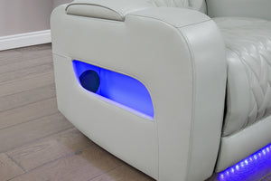 Primer plano del brazo lateral de un sofá reclinable gris claro con tapizado acolchado. Incluye compartimento de almacenamiento abierto con iluminación LED azul integrada y detalle de luz ambiental en la base para un estilo moderno.