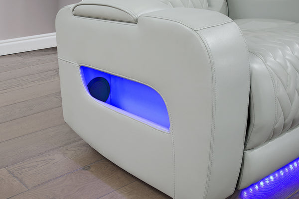 Primer plano del brazo lateral de un sofá reclinable gris claro con tapizado acolchado. Incluye compartimento de almacenamiento abierto con iluminación LED azul integrada y detalle de luz ambiental en la base para un estilo moderno.