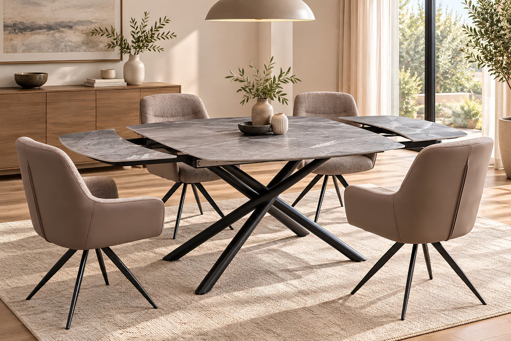 Un juego de comedor moderno que incluye una mesa ovalada de color gris oscuro con efecto piedra y base negra en forma de cruz, rodeada por cuatro sillas tapizadas en beige con patas de metal negro, todo ello sobre una alfombra clara en un comedor luminoso.