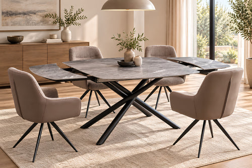 Una configuración de comedor moderno que presenta una mesa rectangular con acabado de piedra y base de metal negro en forma de X, rodeada por cuatro sillas tapizadas en color topo. El mobiliario se dispone sobre una alfombra beige texturizada en un comedor luminoso y contemporáneo, equipado con un aparador de madera, lámparas colgantes y grandes ventanales.