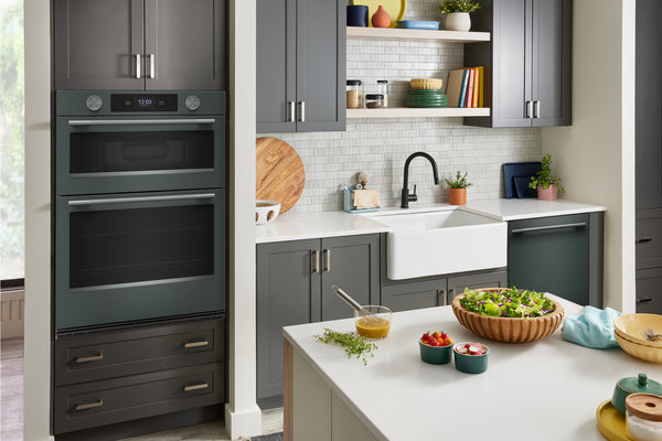 Un interior de cocina moderno que cuenta con un horno doble empotrado en la pared, gabinetes de color verde, encimeras blancas, un fregadero estilo granja, estantes abiertos y alimentos en una isla de cocina.