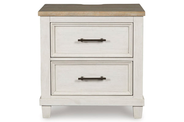 Vista frontal de una mesita de noche antigua de madera blanca con dos gavetas y tiradores negros sobre fondo blanco.