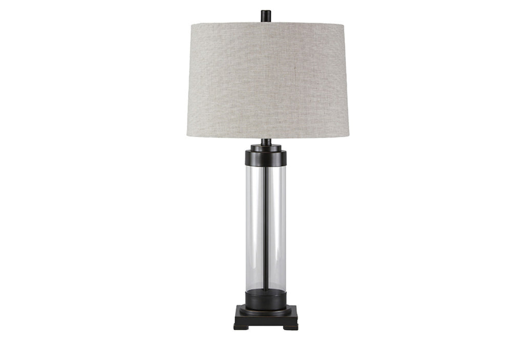 Lámpara de sobremesa con base cilíndrica de cristal y pantalla de tela gris sobre fondo blanco.