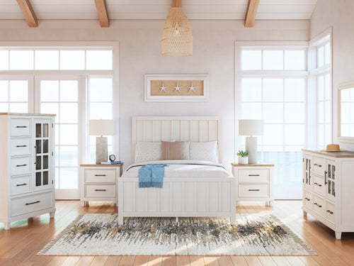 Conjunto de dormitorio antiguo de madera blanca compuesto por una cama, dos mesitas de noche, un chest y un gavetero con espejo, en una habitación luminosa con dos ventanas al fondo.