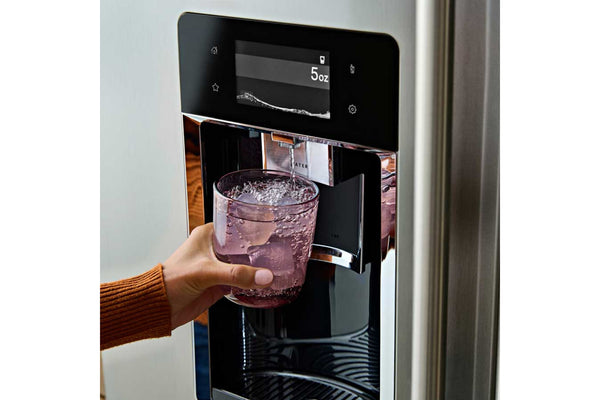 Una mano sostiene un vaso de vidrio transparente debajo de un dispensador de agua negro en un refrigerador de acero inoxidable, con hielo visible en el agua.