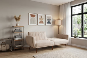 Sofá tipo chaise longue tapizado en tela beige, con respaldo acolchado de líneas verticales, brazos curvos y patas de madera. Presentado en una sala moderna con decoración minimalista, lámpara de pie, estantería y cuadros geométricos en la pared.