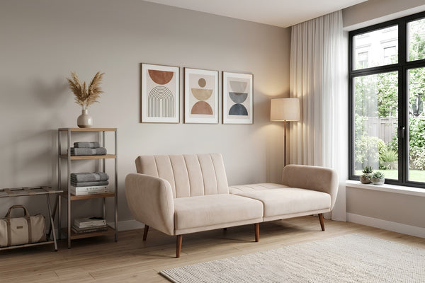 Sofá tipo chaise longue tapizado en tela beige, con respaldo acolchado de líneas verticales, brazos curvos y patas de madera. Presentado en una sala moderna con decoración minimalista, lámpara de pie, estantería y cuadros geométricos en la pared.