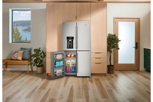 Una nevera de acero inoxidable con congelador superior se muestra en una cocina moderna con gabinetes de madera clara, una encimera, plantas en macetas y vista a la ventana. La puerta del refrigerador está abierta, dejando ver estantes con alimentos.