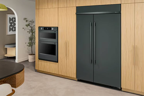 Una escena de cocina moderna que muestra una nevera de dos puertas en color gris oscuro mate integrado en gabinetes de madera clara, con un horno empotrado a juego a su izquierda.