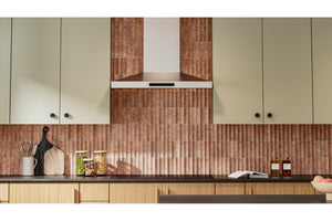 Un interior de cocina moderno que presenta un llamativo salpicadero de azulejos verticales acanalados de cobre o bronce detrás de una encimera oscura, con armarios de madera clara y una campana extractora a juego.