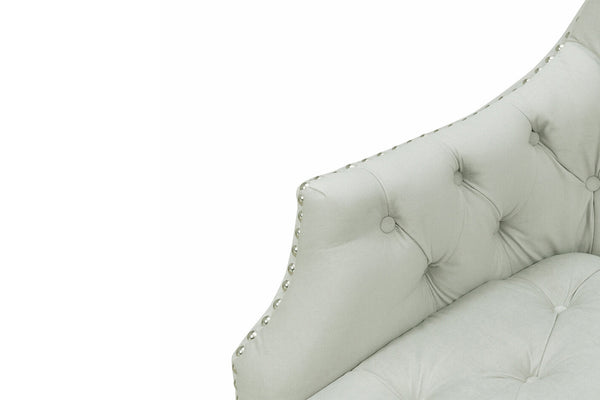 Vista cercana de brazo de silla de acento color crema con respaldo capitonado y detalles de tachuelas, sobre un fondo blanco. 